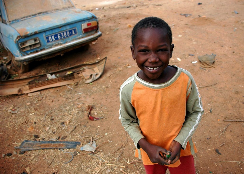 Photograph of a young boy in Senegal, smiling at the camera in front of a broken rusty car
